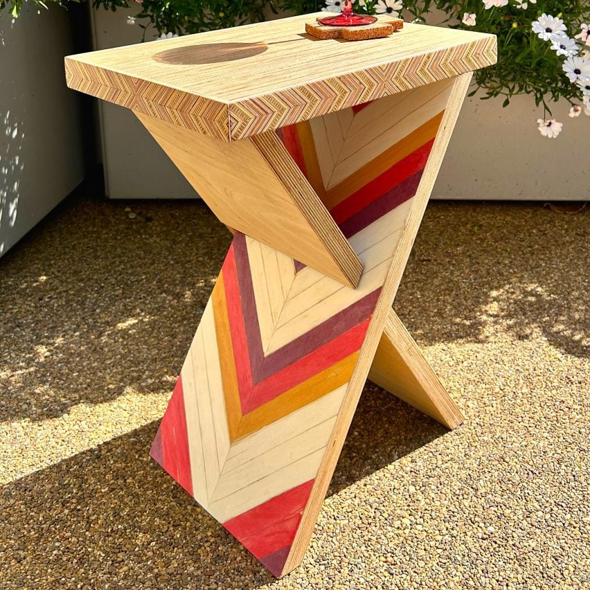 A triangular shaped wooden side table with geometric patterns in red, yellow and brown tones, placed on gravel.